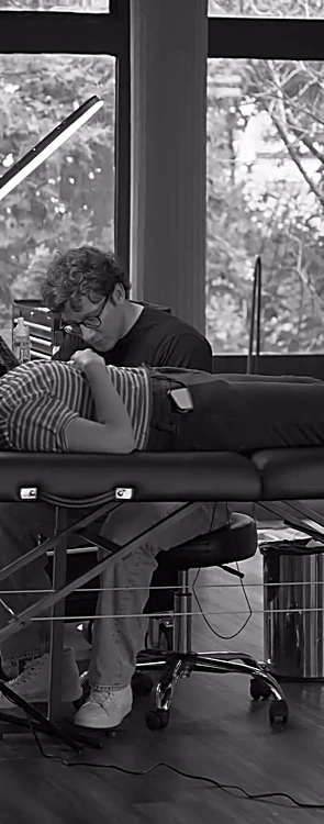 Camilla de tatuaje y artista trabajando frente a los ventanales, en blanco y negro