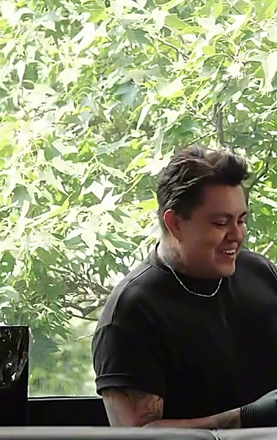 Artista sonriendo en su lugar de trabajo frente a los ventanales, con árboles verdes detrás del vidrio